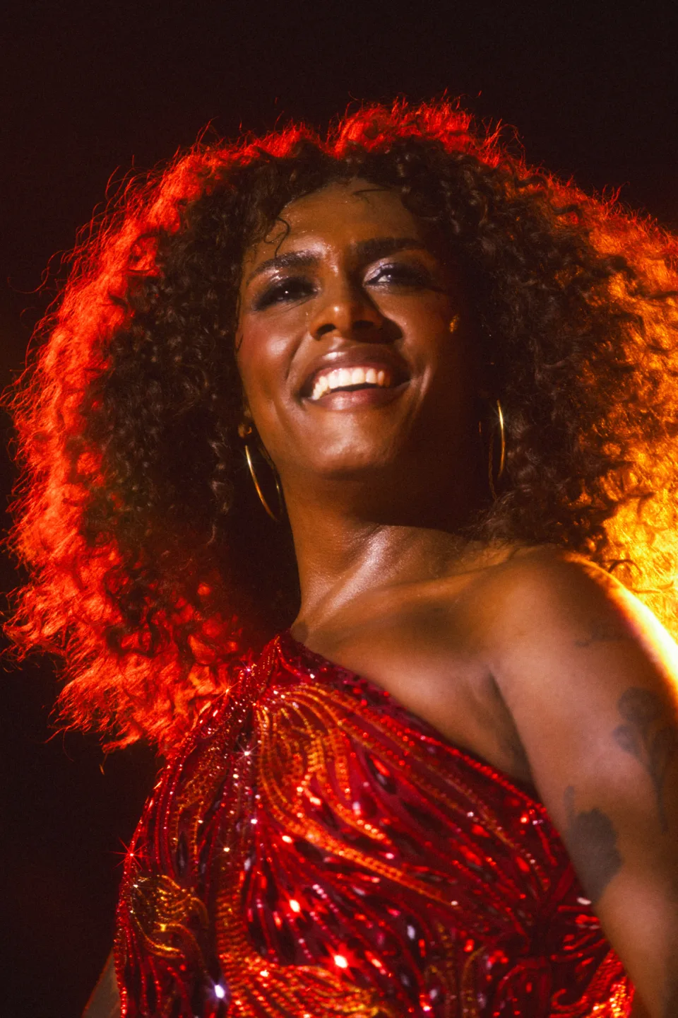 Liniker sorri sob luz vermelha de palco, usando vestido de paetês vermelhos e brincos de argola, durante show da turnê Bye Bye Caju