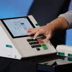Mão pressiona o teclado de uma urna eletrônica da Justiça Eleitoral, com a tela em modo de treinamento, durante apresentação do Tribunal Regional Eleitoral