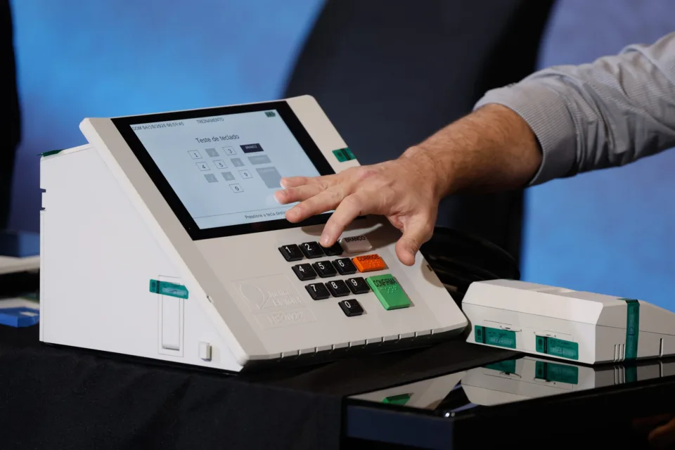 Mão pressiona o teclado de uma urna eletrônica da Justiça Eleitoral, com a tela em modo de treinamento, durante apresentação do Tribunal Regional Eleitoral
