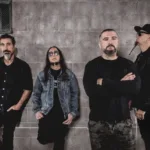 Os quatro integrantes do System of a Down — Serj Tankian, Daron Malakian, John Dolmayan e Shavo Odadjian — posam de preto encostados em uma parede de blocos de concreto