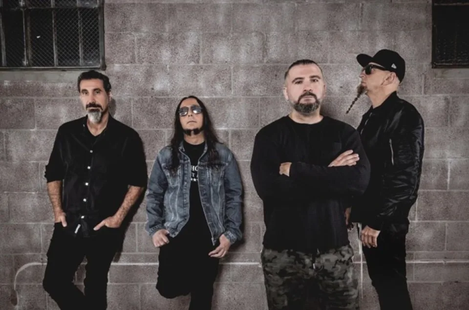 Os quatro integrantes do System of a Down — Serj Tankian, Daron Malakian, John Dolmayan e Shavo Odadjian — posam de preto encostados em uma parede de blocos de concreto