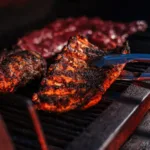 Costela e cortes de carne assando na grelha de uma churrasqueira, com pinça segurando uma das peças, durante festival de churrasco