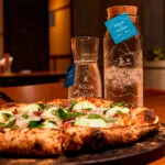 Garrafas de vidro personalizadas do projeto Agua da Casa, da Everest, ao lado de pizza napolitana na Ribalta Pizzaria, no Ribalta Hotel, na Barra da Tijuca