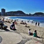 Praia de Copacabana com banhistas, calçadão de mosaico português e prédios da orla ao fundo, no Rio de Janeiro