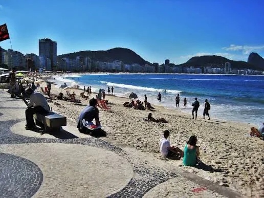 Praia de Copacabana com banhistas, calçadão de mosaico português e prédios da orla ao fundo, no Rio de Janeiro