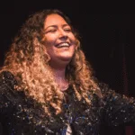 A cantora Amanda Mendonça sorri de braços abertos no palco, com vestido brilhante e fundo escuro