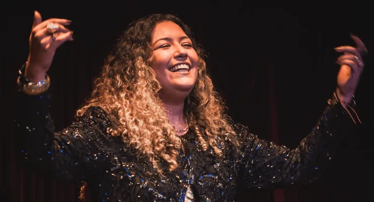 A cantora Amanda Mendonça sorri de braços abertos no palco, com vestido brilhante e fundo escuro