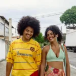 Duas jovens sorridentes caminham por uma rua de subúrbio em imagem de divulgação do Cineclube Curta o Curta