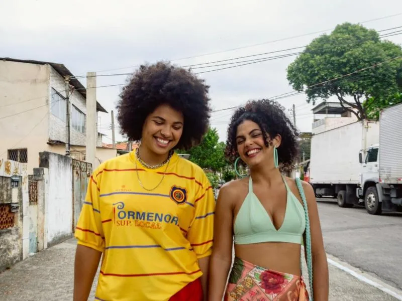 Duas jovens sorridentes caminham por uma rua de subúrbio em imagem de divulgação do Cineclube Curta o Curta