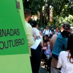 Crianças e adultos reunidos sob as árvores do Parque Lage ao lado de um painel verde com o título Jornadas de Outubro