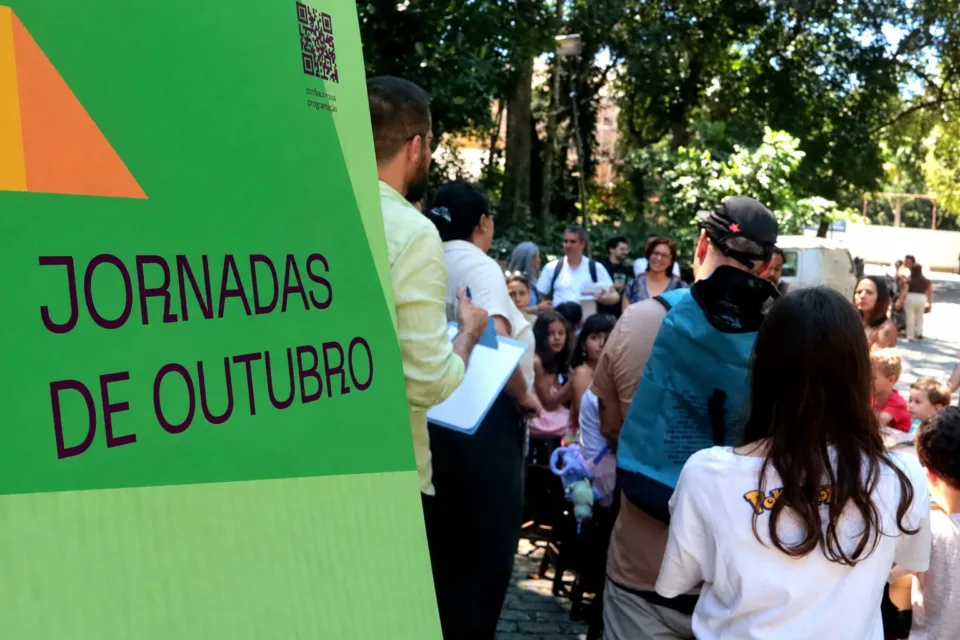 Crianças e adultos reunidos sob as árvores do Parque Lage ao lado de um painel verde com o título Jornadas de Outubro