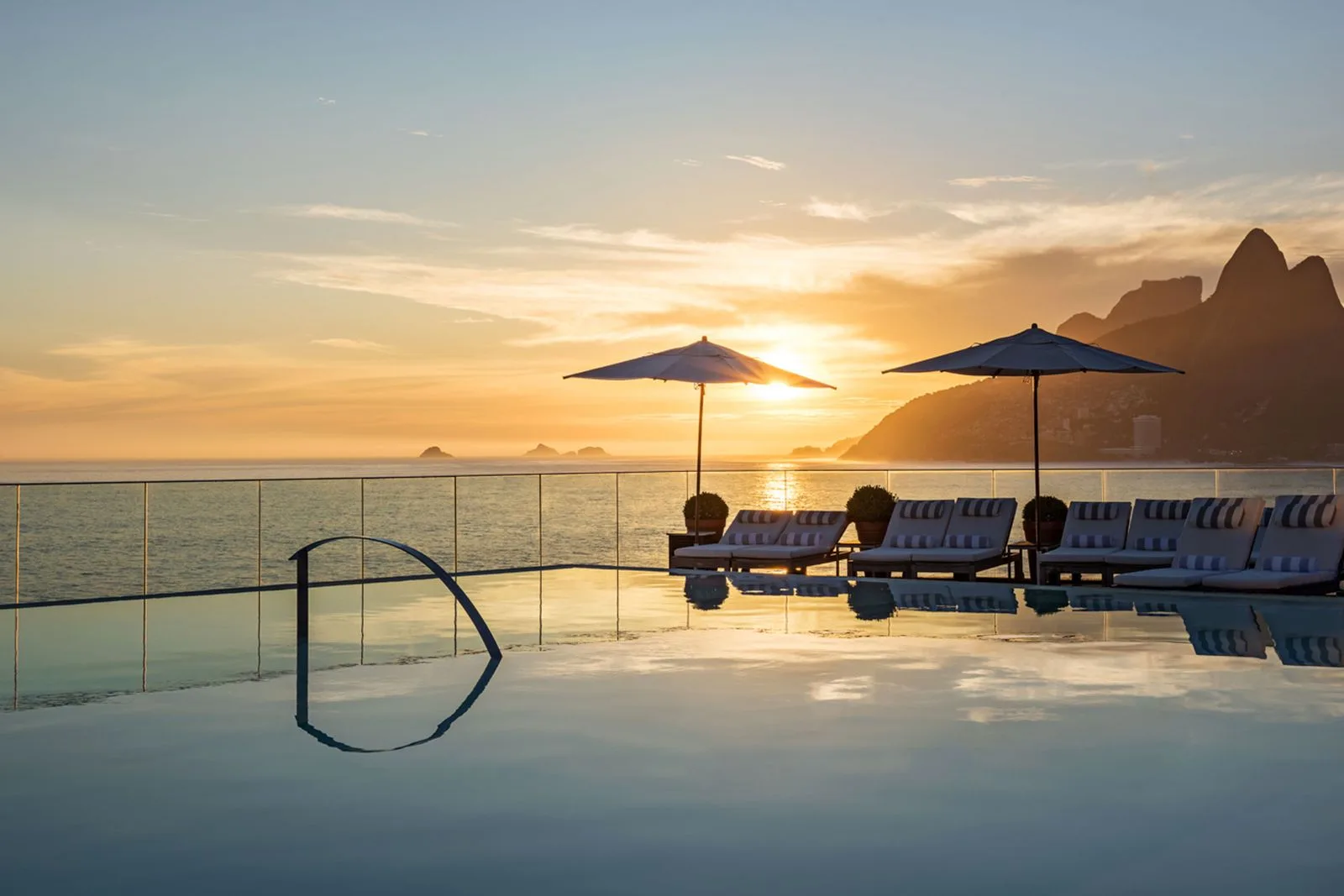 Piscina de borda infinita no rooftop do Hotel Fasano Rio de Janeiro ao pôr do sol, com guarda-sóis e espreguiçadeiras, o mar de Ipanema e o Morro Dois Irmãos ao fundo