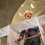 Ator caracterizado como Santos Dumont, de chapéu e bigode, segura estrutura de bambu e tecido branco que remete ao 14-Bis