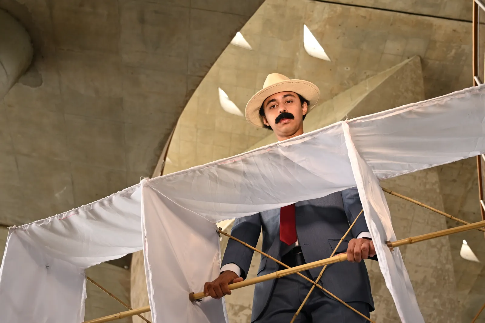Ator caracterizado como Santos Dumont, de chapéu e bigode, segura estrutura de bambu e tecido branco que remete ao 14-Bis