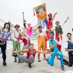 Integrantes da Orquestra Circônica, com figurinos coloridos e instrumentos de sopro e percussão, posam com o estandarte do grupo