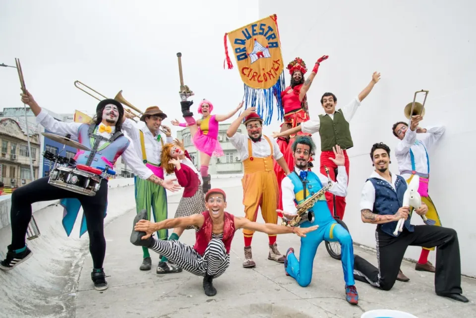 Integrantes da Orquestra Circônica, com figurinos coloridos e instrumentos de sopro e percussão, posam com o estandarte do grupo