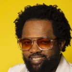 Retrato do sambista Xande de Pilares, de óculos e paletó claro, sobre fundo amarelo