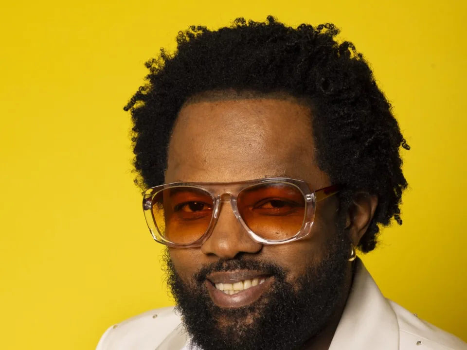 Retrato do sambista Xande de Pilares, de óculos e paletó claro, sobre fundo amarelo